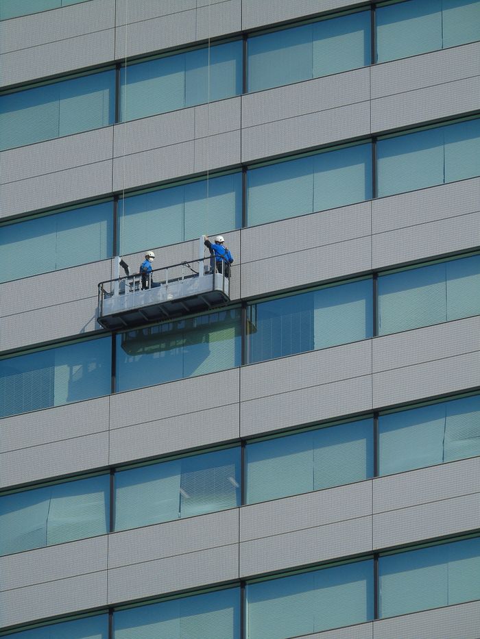 Deux ouvriers nettoient la façade vitrée d’un bâtiment moderne à l’aide d’une plateforme suspendue.
